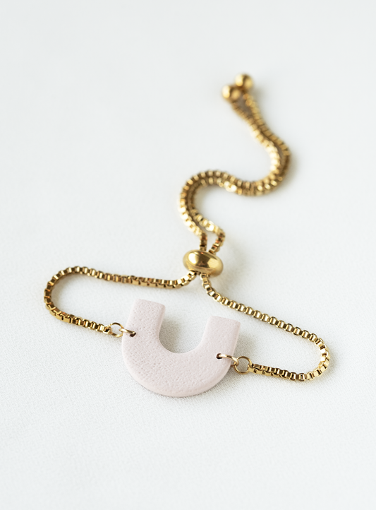 adjustable gold plated stainless steel bracelet with a pale pink arch pendant on a white background